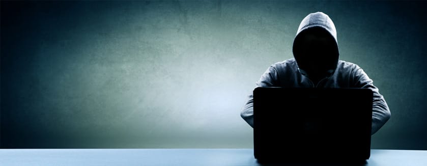 Ein Mann im Kapuzenpullover sitzt mit einem Laptop an einem Schreibtisch und untersucht Sicherheitslücken.