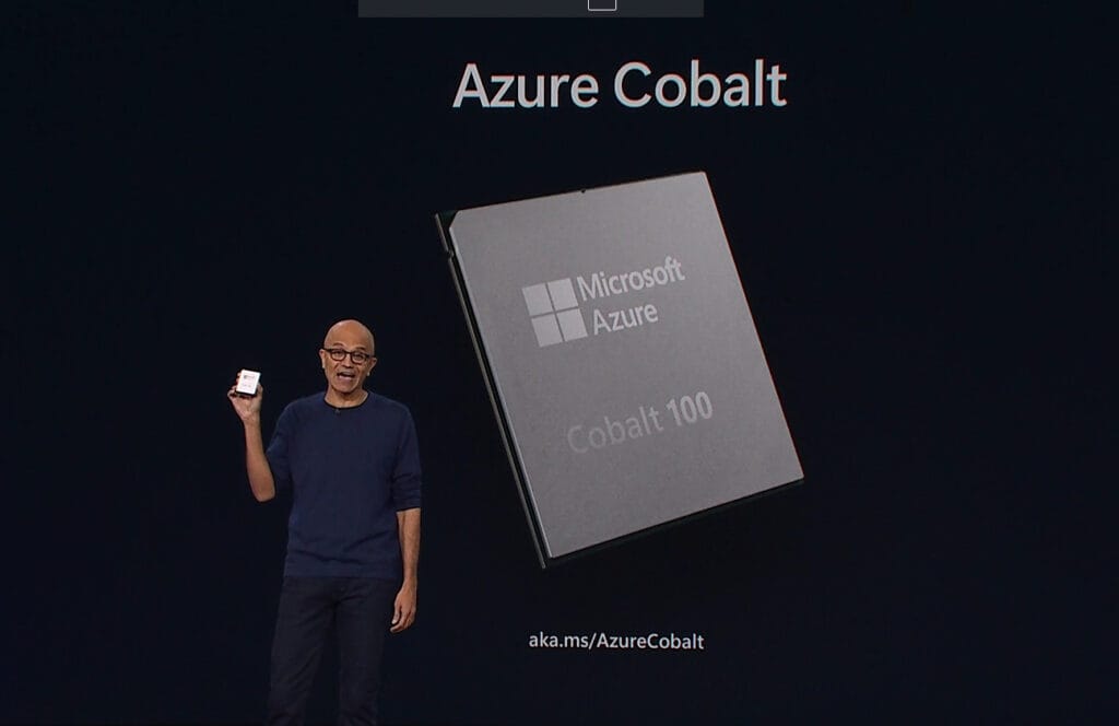 Satya Nadella hält den silbernen Azure Cobalt 100 CPU in der Hand. Hinter ihm auf der grossen Leinwand ist eine Nahaufnahme des Chips eingeblendet